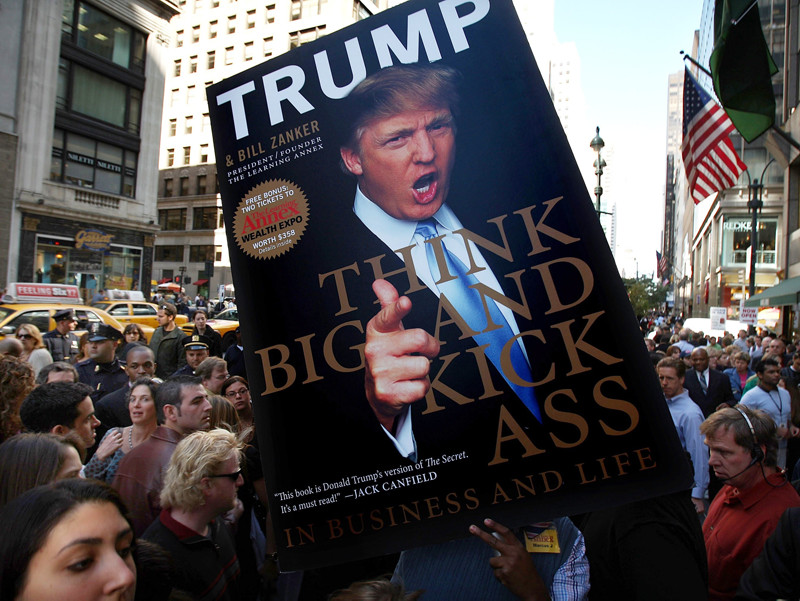 Photo of a person holding a life-size version of the cover of Donald Trump's book, Think Big and Kick Ass In Business and Life, among a crowd of others.